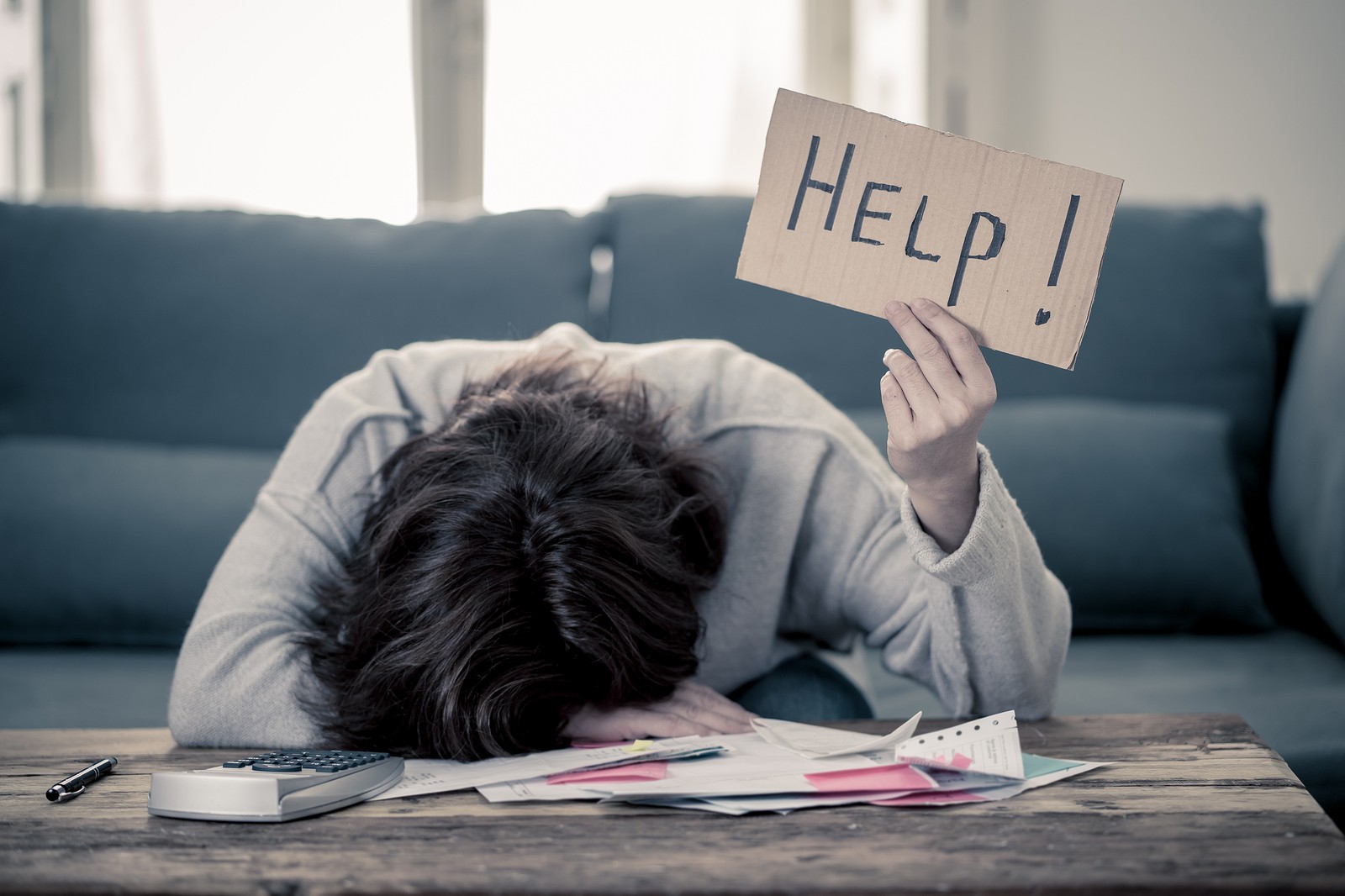 Person with head down on a desk covered in papers, looking distraught. Holding a paper, handmade sign that says "Help!". This person could definitely benefit from Portland-area counseling for life transitions.