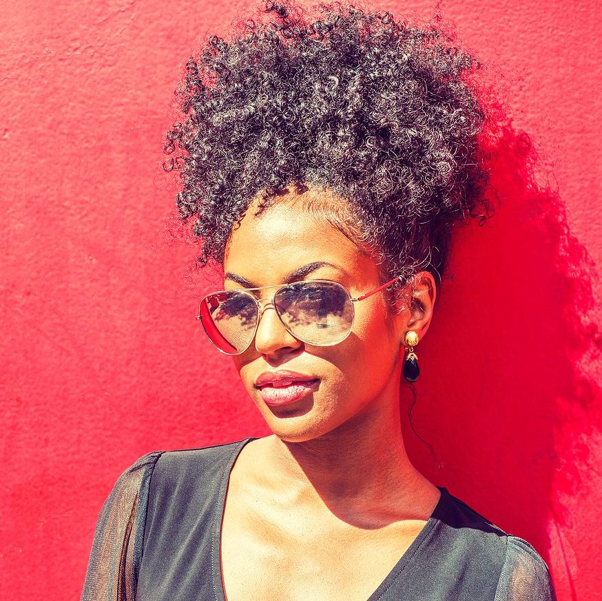 Young black woman with afro hairstyle wearing long sleeve mesh sheer shirt, sunglasses, standing against red background under sun. Meeting with an online therapist in Oregon is meeting with another human, just like you!
