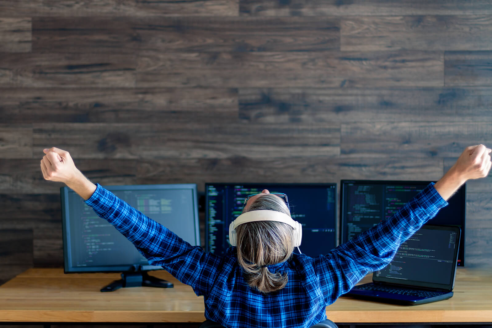 A person wearing a blue plaid shirt and headphones raises their arms in triumph while sitting at a desk with multiple computer monitors displaying code. This could represent feeling more fulfilled at work after finding balance through therapy in Tigard, OR. Search for online therapy in Oregon and how an Oregon online therapist can help today. 
