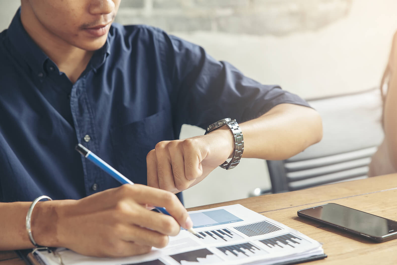 A man in a navy blue shirt sits at a table, writing on a document with graphs while glancing at his watch. This could symbolize the balance between work and personal time. Contact an online therapist in Oregon to learn more about the benefits of therapy in Tigard, OR and anxiety therapy can help address workplace issues.
