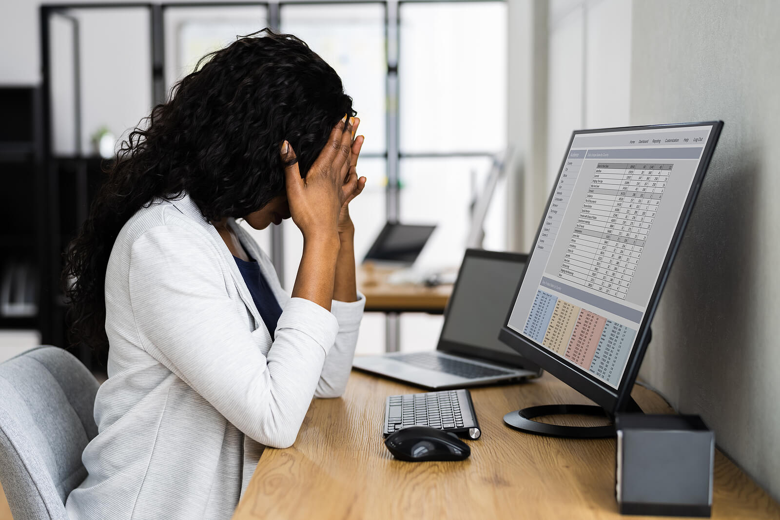 A woman in a white blazer sits at a desk with her hands covering her face, appearing overwhelmed while looking at a computer screen displaying data. Search for how an online therapist in Oregon can offer support with anxiety therapy in Tigard, OR. Search for a Christian therapist in Tigard, OR for more support today.
