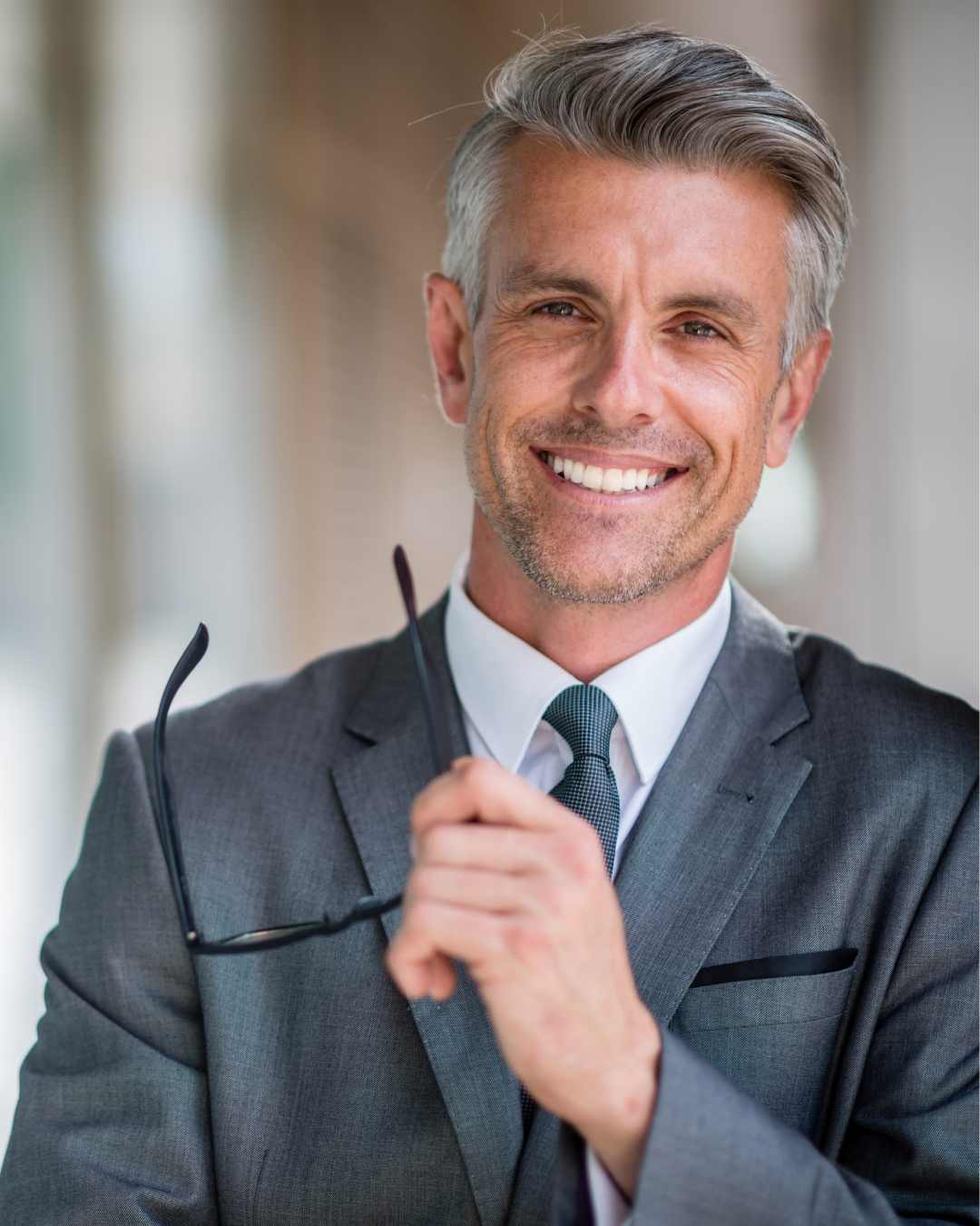 A middle-aged man in a grey suit smiles while holding a pair of eyeglasses in his hand. Perfectionism in men in Tigard, OR can be helped with therapy.