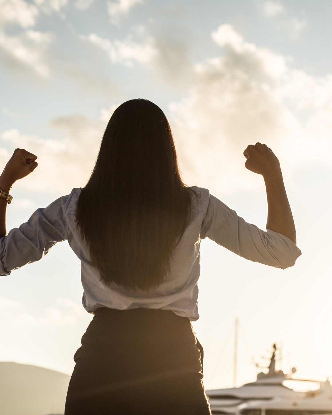 A woman stands outdoors with her back to the camera, raising both fists in the air against a backdrop of sky and clouds.