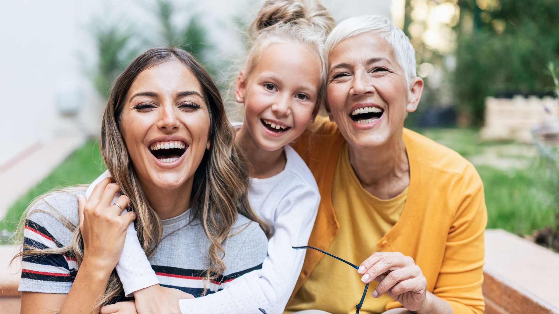 Three women of different ages, likely from three generations, sit close together outdoors, smiling and laughing at the camera. Perfectionism in women in Tigard, OR can be helped with therapy.