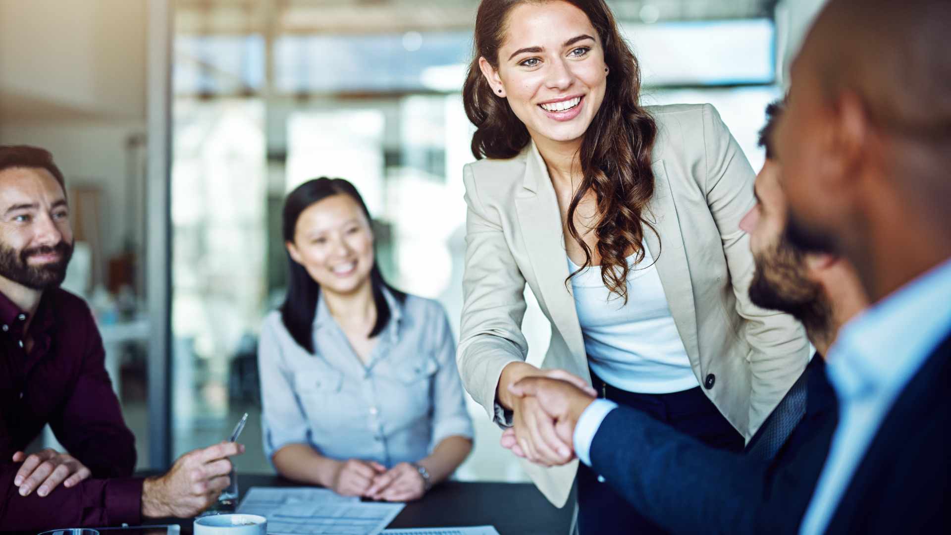 A woman in business attire shakes hands with a seated man during a meeting, while two other colleagues look on and smile in a modern office setting.
