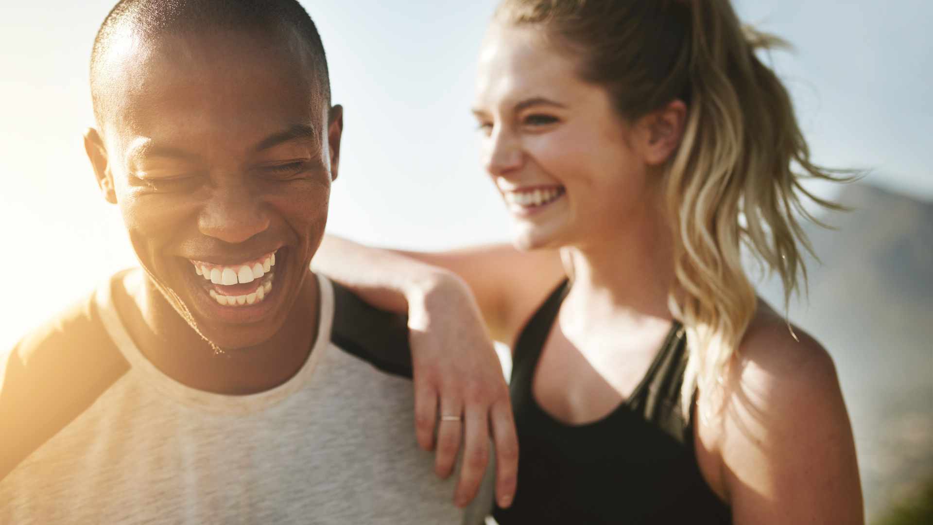 Two people standing close together outdoors, smiling and laughing, with one person’s arm resting on the other’s shoulder.