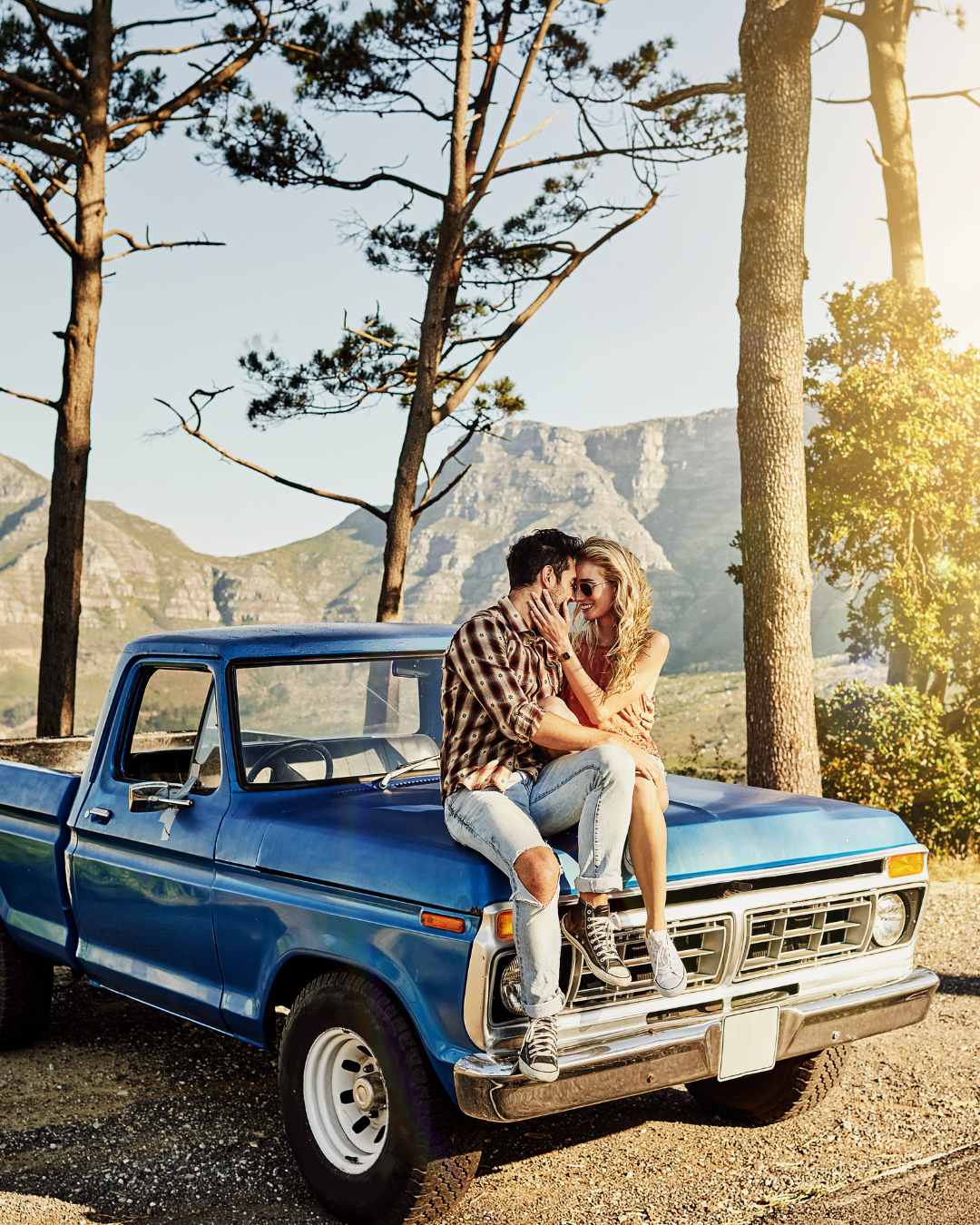 A man and woman sit close together on the hood of a blue vintage pickup truck, surrounded by trees and mountains in the background.