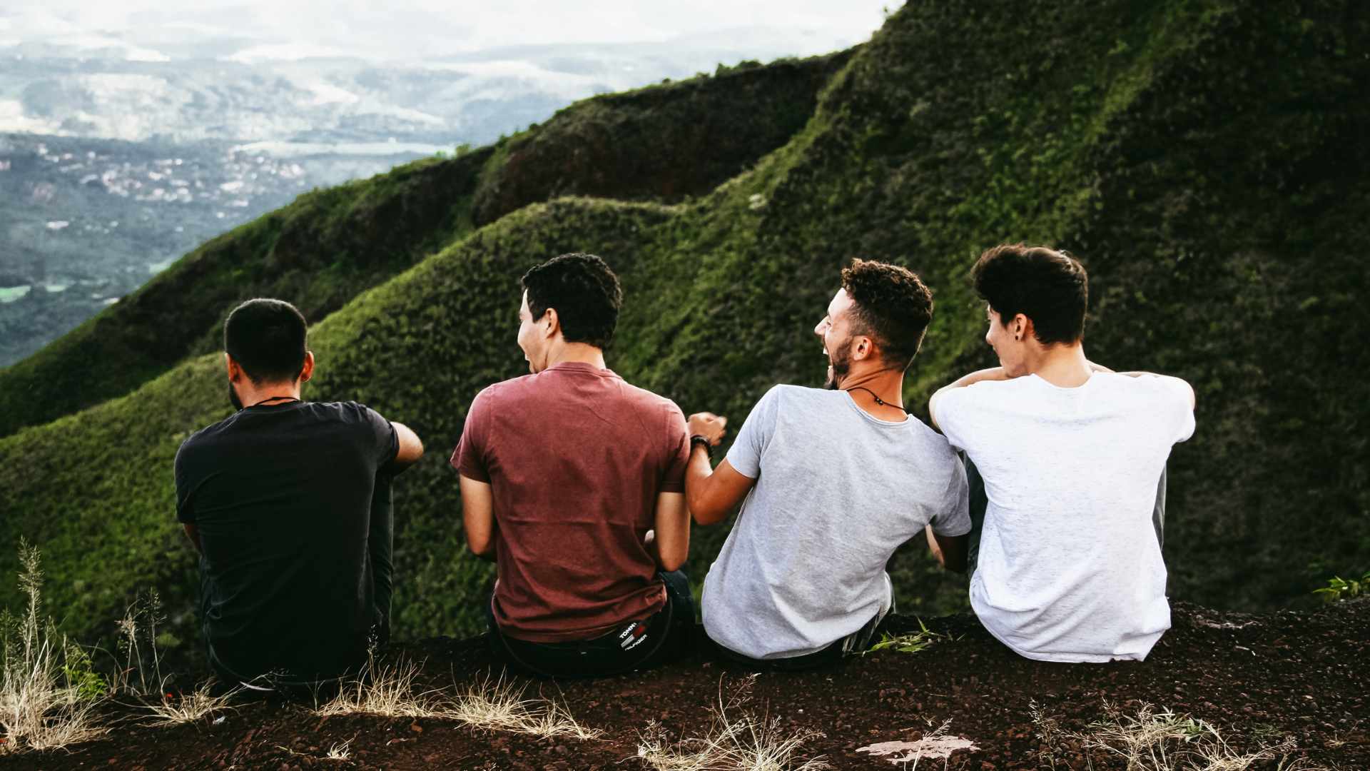 Four young men sit on a grassy hillside facing away from the camera, overlooking green mountains and valleys in the background.