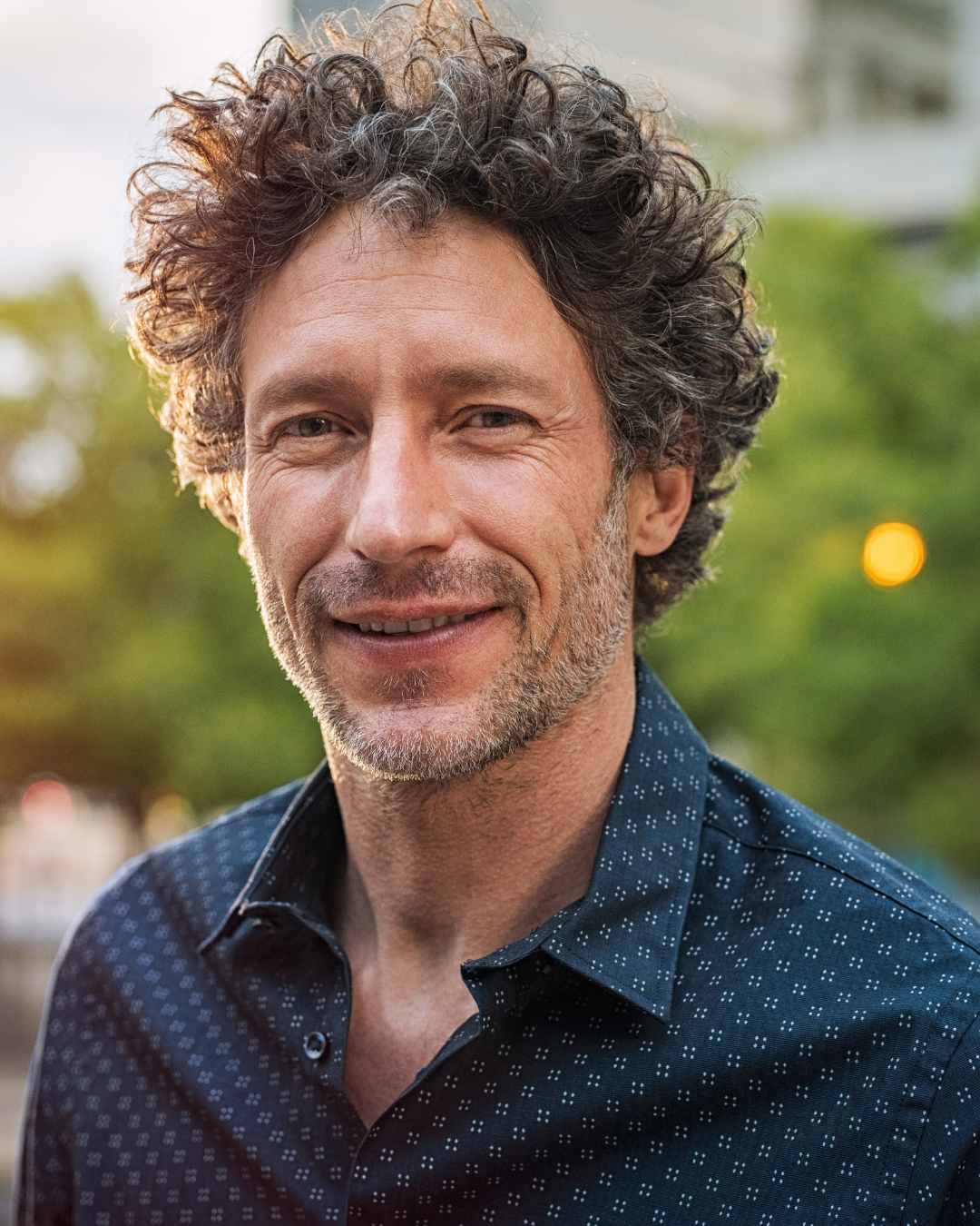 Man with curly hair and a beard wearing a dark patterned shirt, smiling outdoors with greenery and blurred lights in the background.