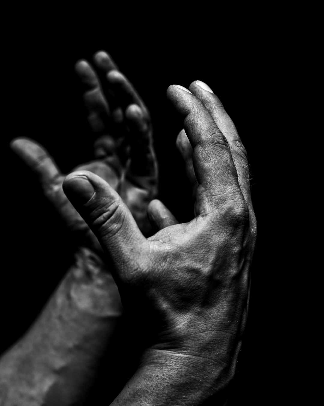 A black and white photo showing two rough, outstretched hands with visible veins and wrinkles against a dark background.