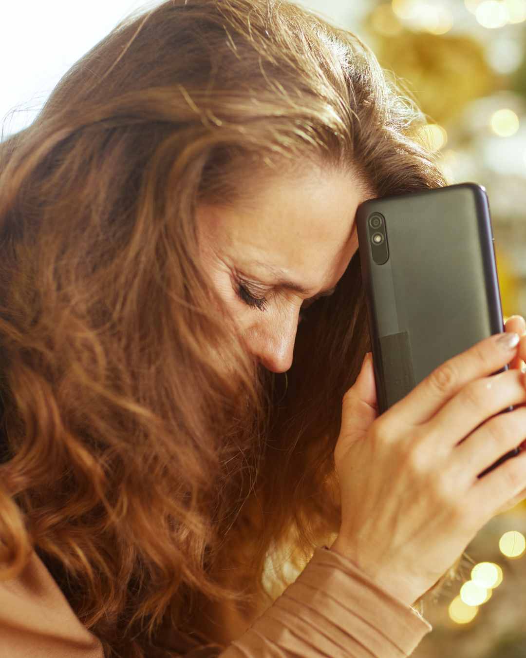 A woman with long brown hair holds a smartphone to her forehead, eyes closed, appearing distressed or upset.