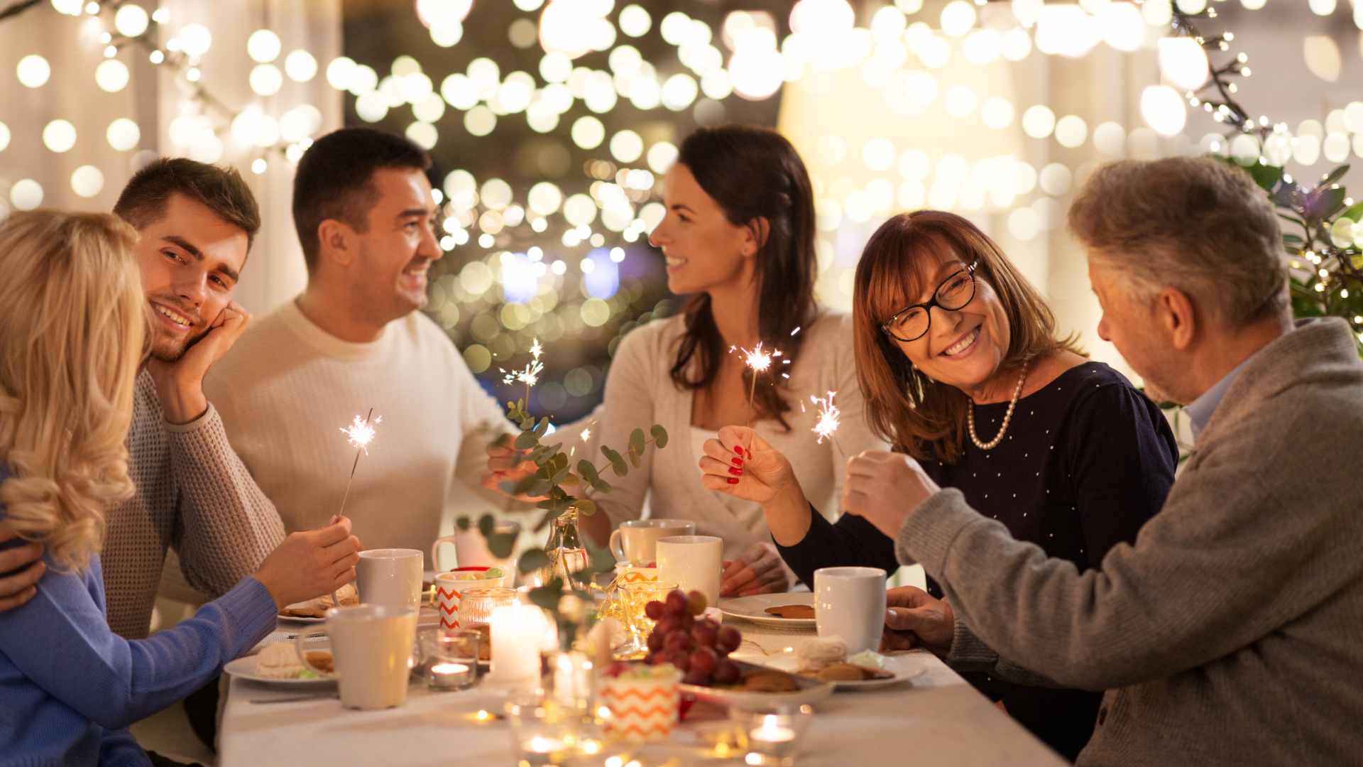A group of people sit around a decorated table, holding sparklers and smiling, with string lights in the background.