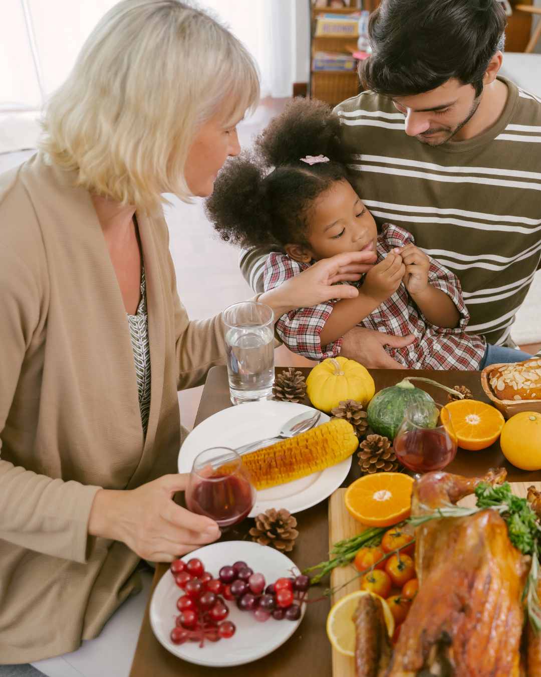 Three people sit at a table with various foods, including corn, oranges, grapes, and a roasted dish, sharing a meal together.
