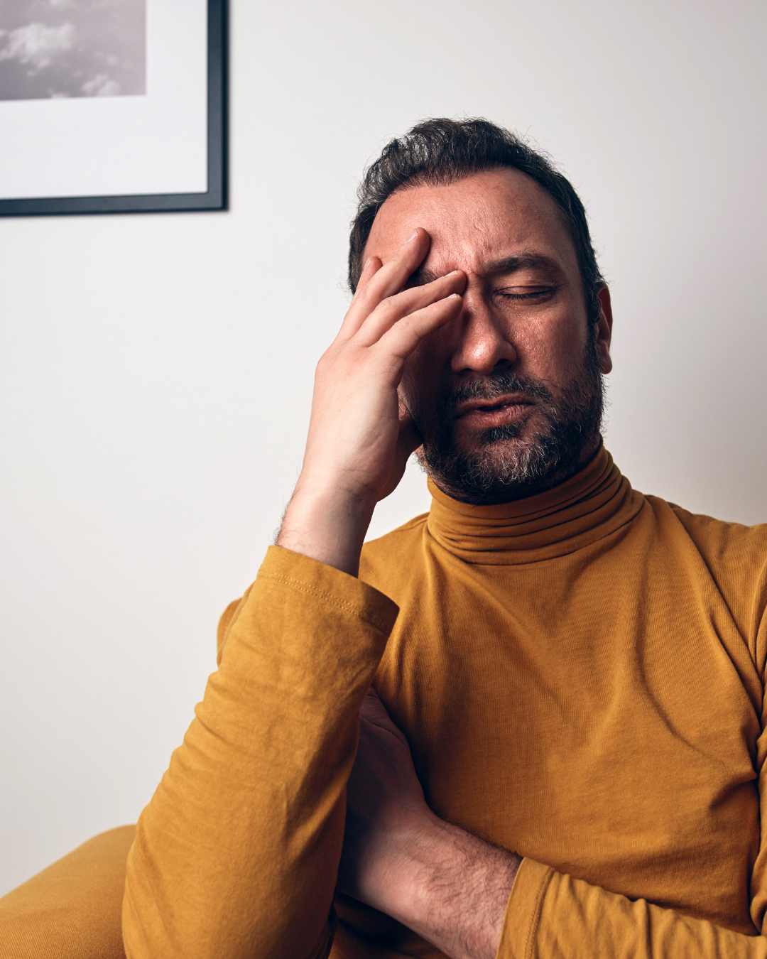 Stressed Man | Overthinking and Perfectionism in Tigard, OR A man in a mustard turtleneck sits with eyes closed and his hand on his forehead, appearing stressed or fatigued from overthinking and perfectionism in Tigard, OR