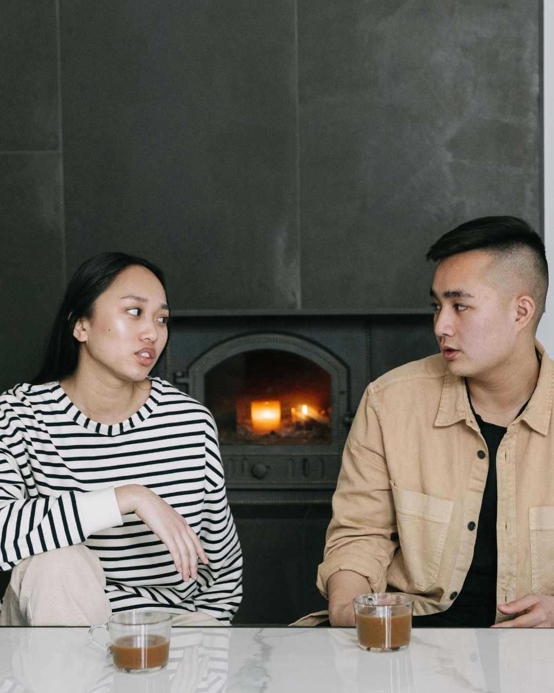 Two people are sitting at a table with drinks, sharing a moment of vulnerability as they converse in front of a lit fireplace. Being vulnerable in relationships can help with intimacy.
