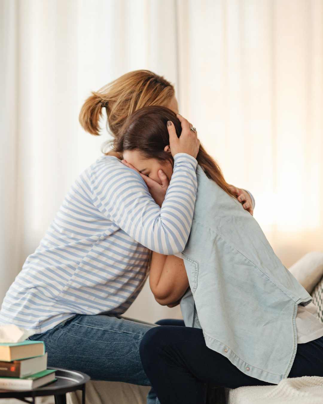 Two women sit on a couch hugging each other closely, one comforting the other in a moment of vulnerability. A small stack of books is visible on the table beside them. Vulnerability can help with intimacy. Get help with this from a Tigard, OR therapist.