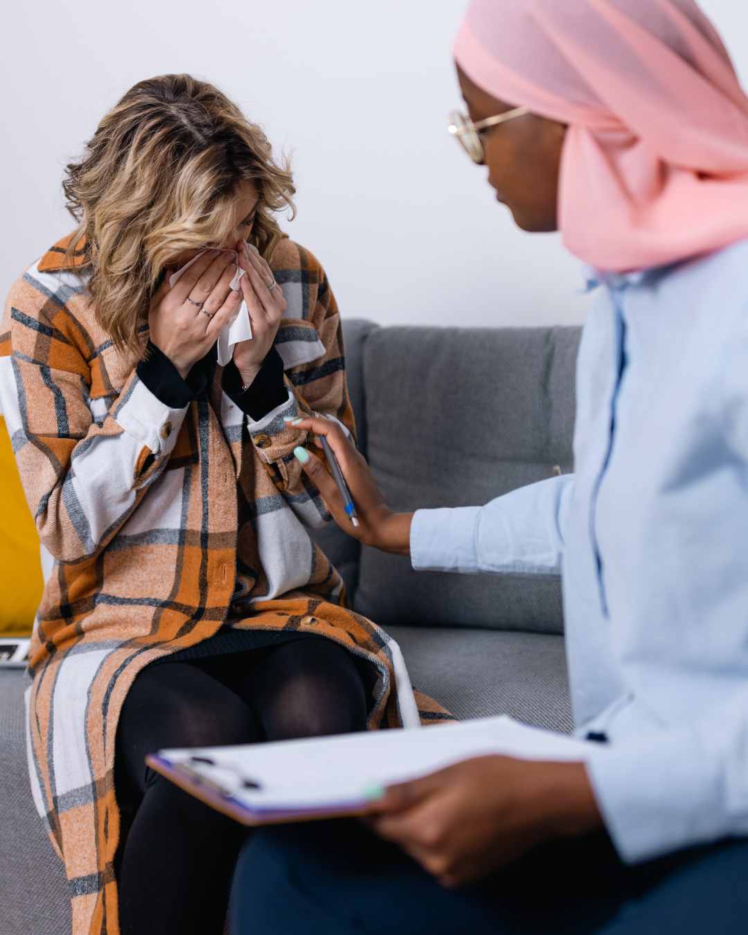 A woman sits on a couch covering her face with her hands while another woman, wearing a pink headscarf, offers comfort and holds a clipboard.